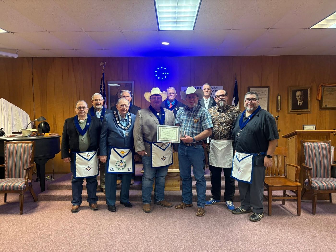 Lodge members in white aprons stand in the lodge room with Caldwell County Sheriff Mike Lane, who holds a Community Builder Award certificate and trophy
